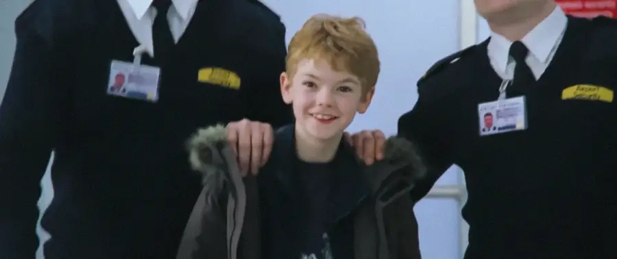 A smiling young boy with red hair stands between two airport security officers in uniform, with their hands resting on his shoulders.
