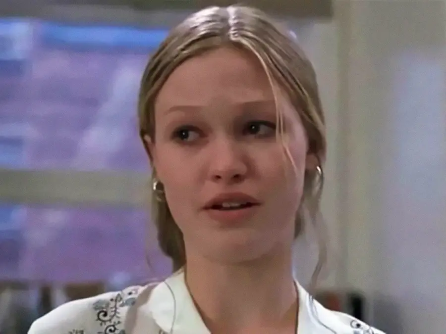 A young woman with blonde hair, parted in the middle and tied back, looks emotional with teary eyes. She is indoors, wearing a light-colored top and hoop earrings, with a blurred window in the background.
