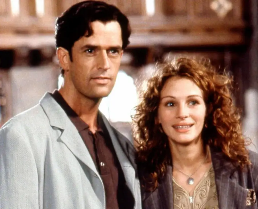A man with dark hair in a light-colored jacket stands next to a smiling woman with wavy hair and braces. They are indoors, with a softly blurred background.