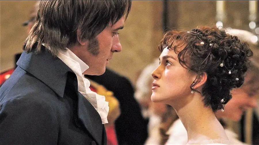 A man in a dark coat and a woman with curly hair and pearls look intently at each other at a formal event, dressed in historical attire from the early 19th century.