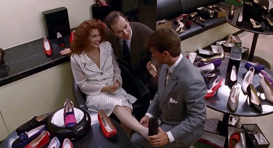A smiling woman in a striped dress sits while a man in a suit helps her try on shoes in a store, with another man beside her and various shoes displayed around them.