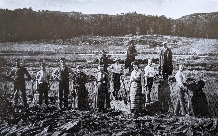 A group of men, women, and children stand in a row outdoors on rocky ground, with hills and trees in the background, dressed in early 20th-century rural clothing.