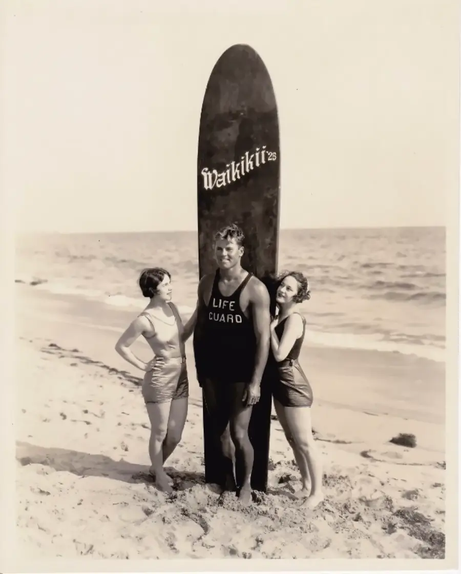 Three people in swimsuits pose on a sandy beach. A man wearing a "LIFE GUARD" tank stands in front of a tall surfboard labeled "Waikiki," flanked by two women. The ocean is in the background.
