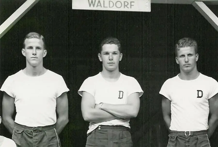 Three young men stand in front of a sign reading "Waldorf." They wear white t-shirts with a letter "D" and dark pants, posing in a row and looking at the camera.