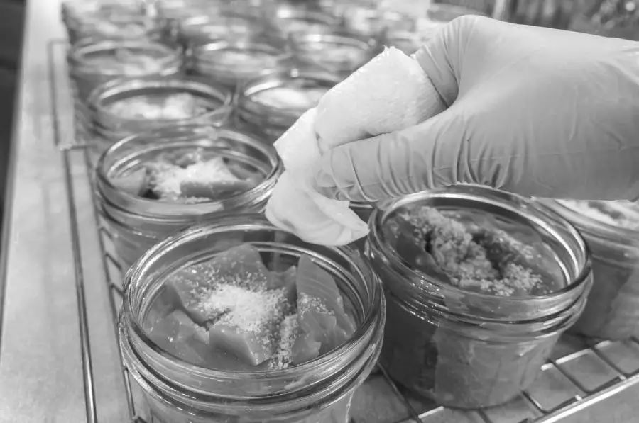 A gloved hand wipes the rim of a glass jar filled with chopped tomatoes and salt; several similar jars sit on a cooling rack in the background.