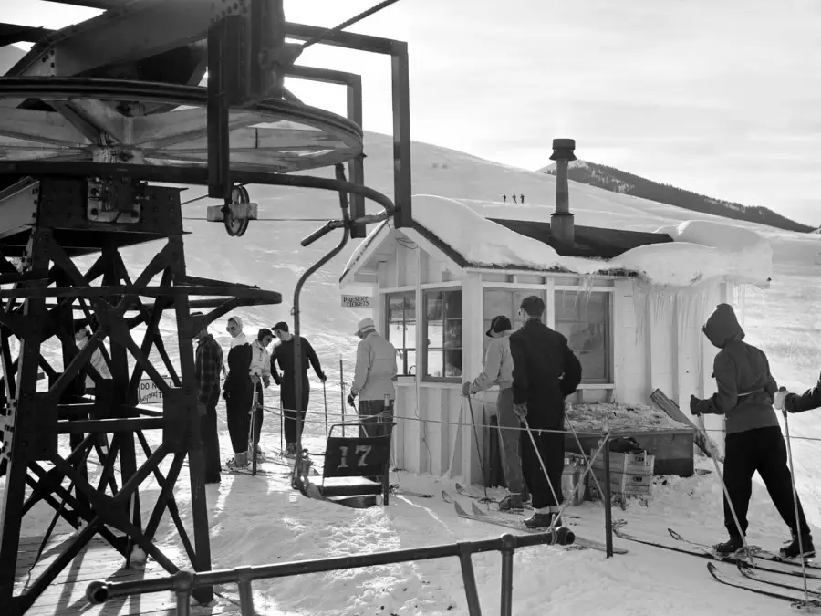 Skiers wait in line at a snowy ski lift station near a small cabin. Snow covers the ground and roof, and a large metal ski lift structure is visible on the left. People are wearing winter clothing and skis.