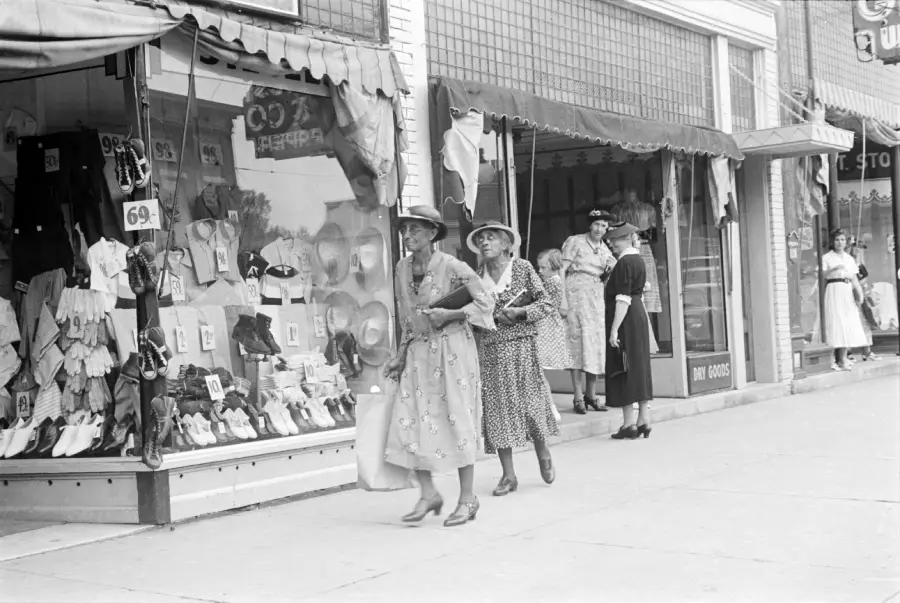 Two women in dresses and hats walk past a store window displaying clothing and accessories. Other women stand and look at the shop window on a busy sidewalk in front of multiple storefronts.