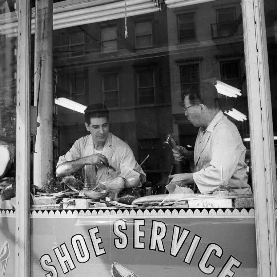 Two men work on repairing shoes behind a shop window that reads "Shoe Service." Tools and shoes are spread across their workbench inside the shop.