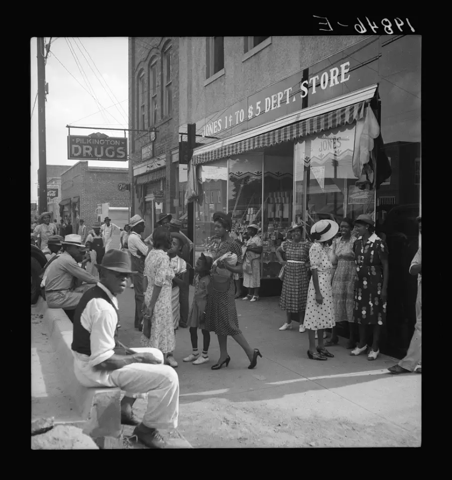 Black and white photo of people gathered in front of Jones 10 to 5$ Dept. Store. Women and children in dresses stand and talk, while a few men sit on benches. A Pilkington Drugs sign is visible in the background.