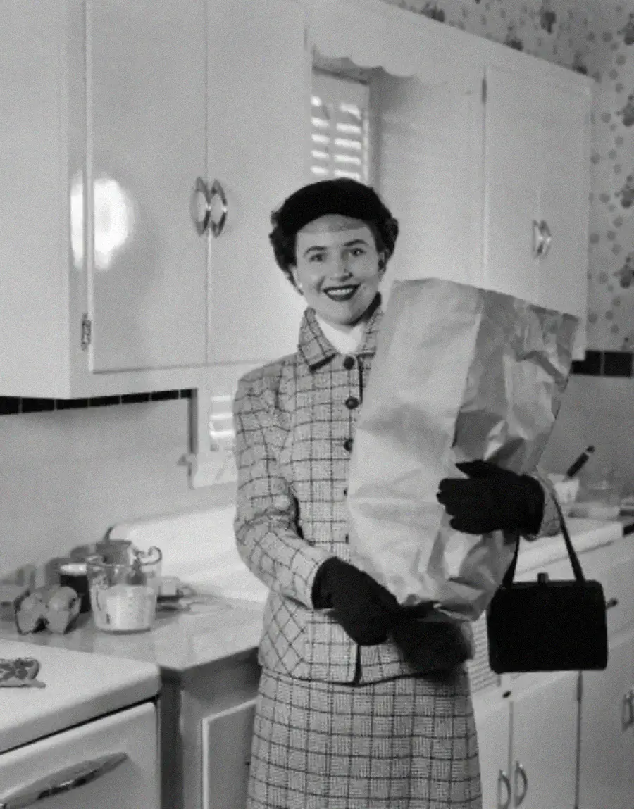 A woman in a plaid coat and gloves smiles while holding a large grocery bag in a vintage kitchen setting.
