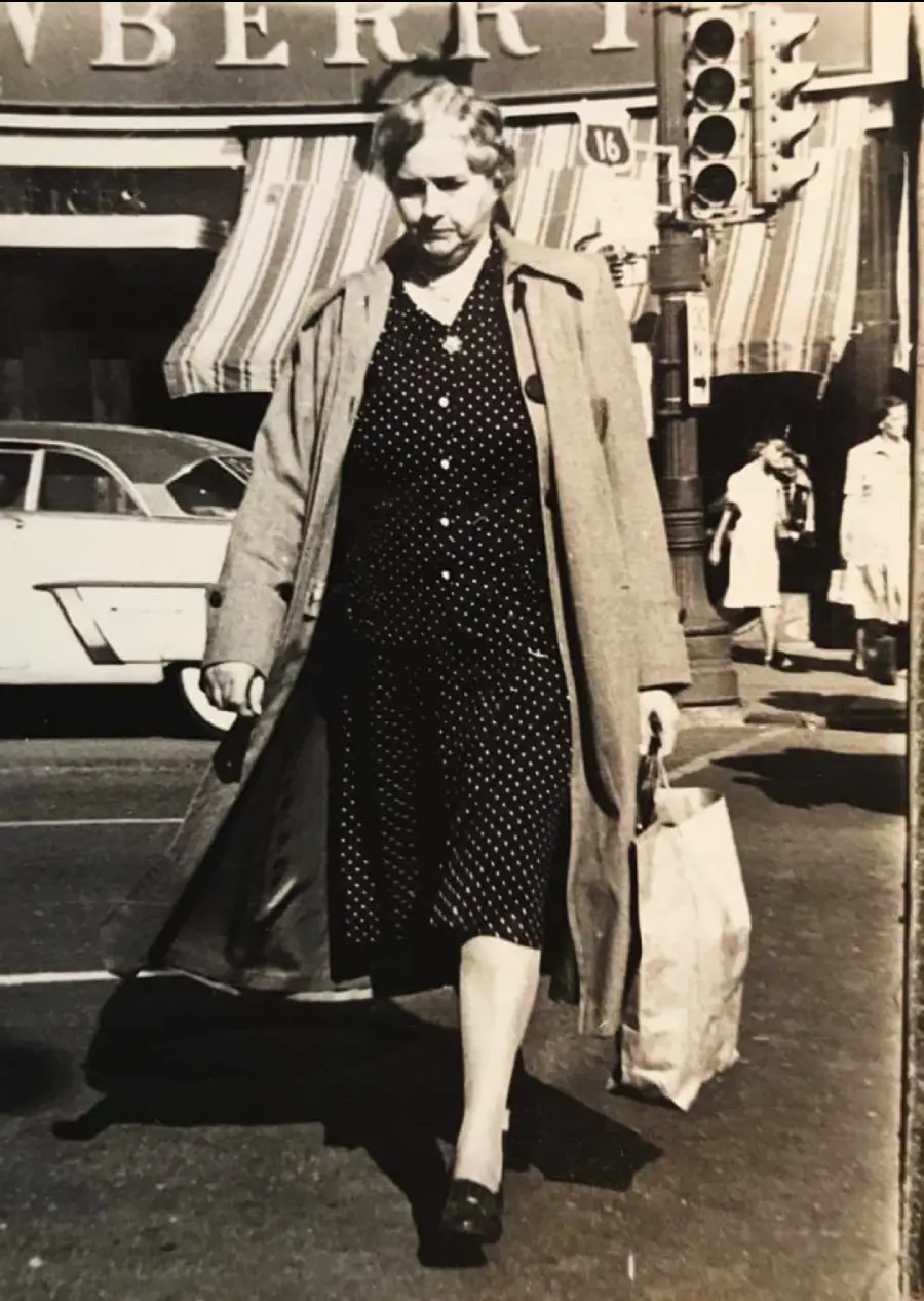 A woman in a polka dot dress and long coat walks across a city street carrying a paper bag. Vintage cars and pedestrians are visible in the background.
