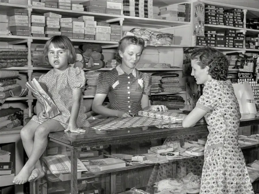 A young girl sits on a counter holding fabric as a woman behind the counter helps another woman choose fabric in a store filled with neatly stacked textiles and sewing supplies.