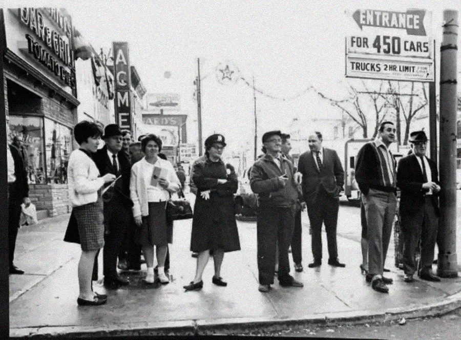 A group of people stand on a city sidewalk near a parking lot entrance; vintage cars, store signs, and 1960s-era clothing are visible in the black-and-white photo.