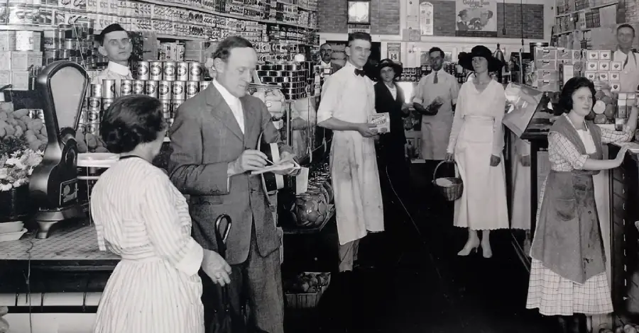 Black and white photo of a vintage grocery store with men and women shopping and working; shelves are filled with canned goods, and people are dressed in early 20th-century clothing.