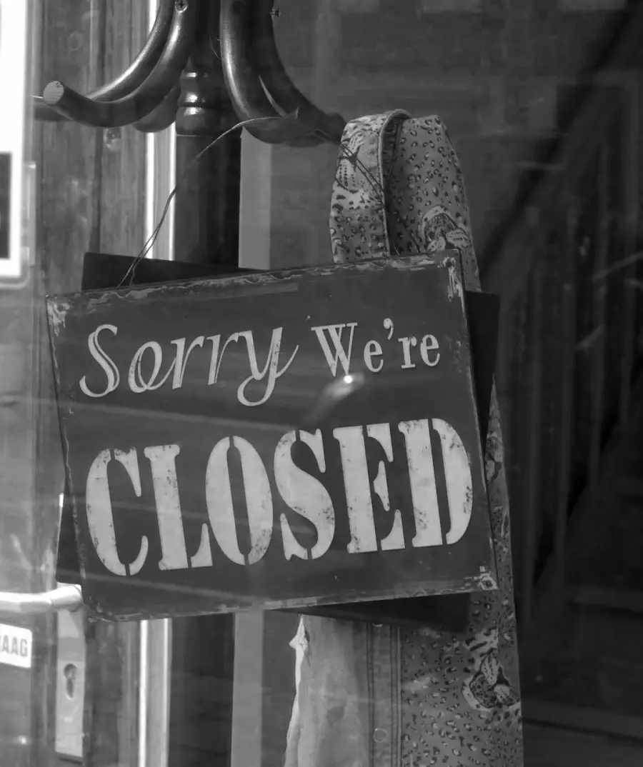 A black and white photo of a sign hanging on a door handle that reads, "Sorry We're CLOSED." A patterned cloth or scarf is draped next to the sign.