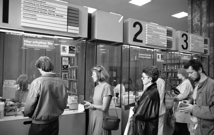 People stand in line at counters inside a bank or post office. Signs above the counters provide information and numbers. The image is black and white, with customers holding documents or bags.