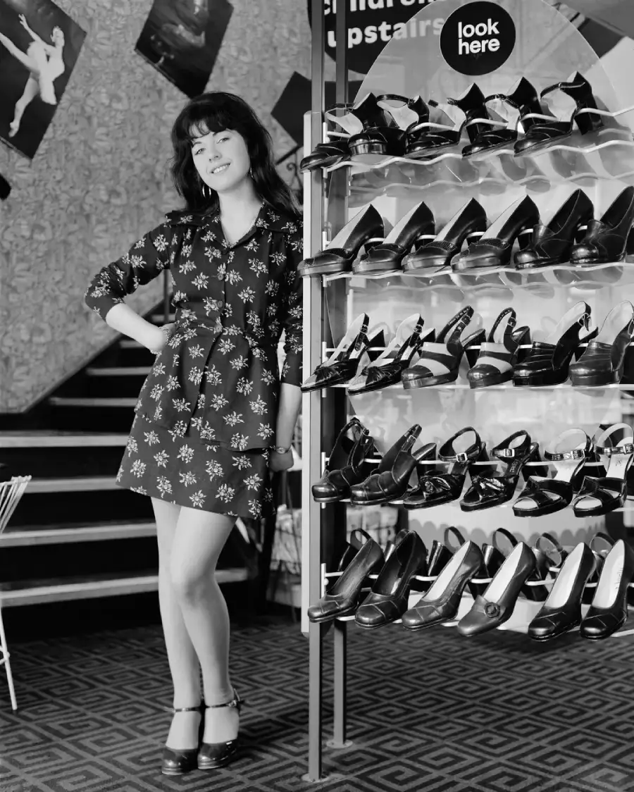 A woman in a patterned dress stands smiling beside a shoe display filled with high heels. She poses with one hand on her hip. Stairs and posters are visible in the background. The photo is black and white.
