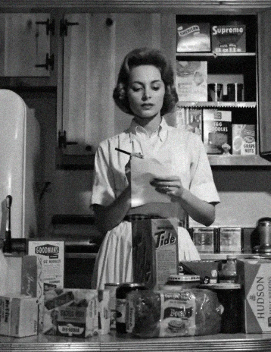 A woman in a vintage kitchen reads a piece of paper, surrounded by boxes of groceries and household products on the counter. Shelves behind her are stocked with pantry items. The image is black and white.