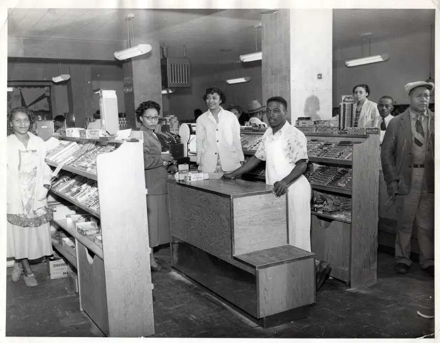 Black-and-white photo of people in a grocery or drugstore. Several employees stand behind the counter, while others stand in aisles looking at the camera. Store shelves are stocked with various goods.
