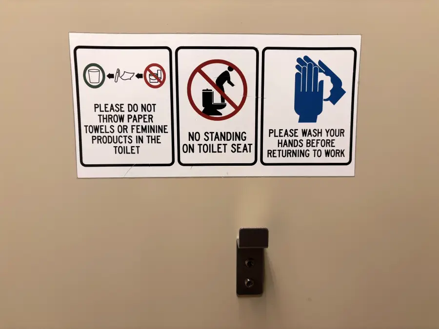 A sign on a bathroom stall door has three notices: do not throw paper towels or feminine products in the toilet, no standing on the toilet seat, and wash hands before returning to work.