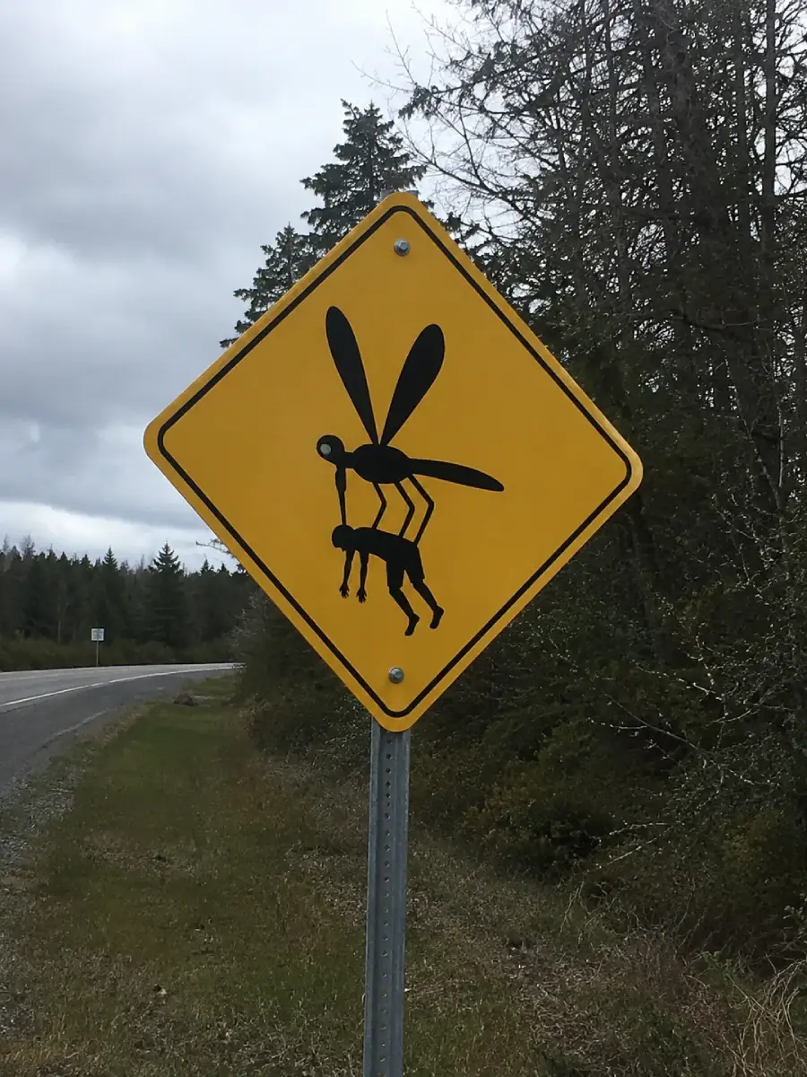 A yellow road sign depicts a large black insect resembling a wasp carrying a deer, set against a backdrop of trees and an overcast sky along a rural road.