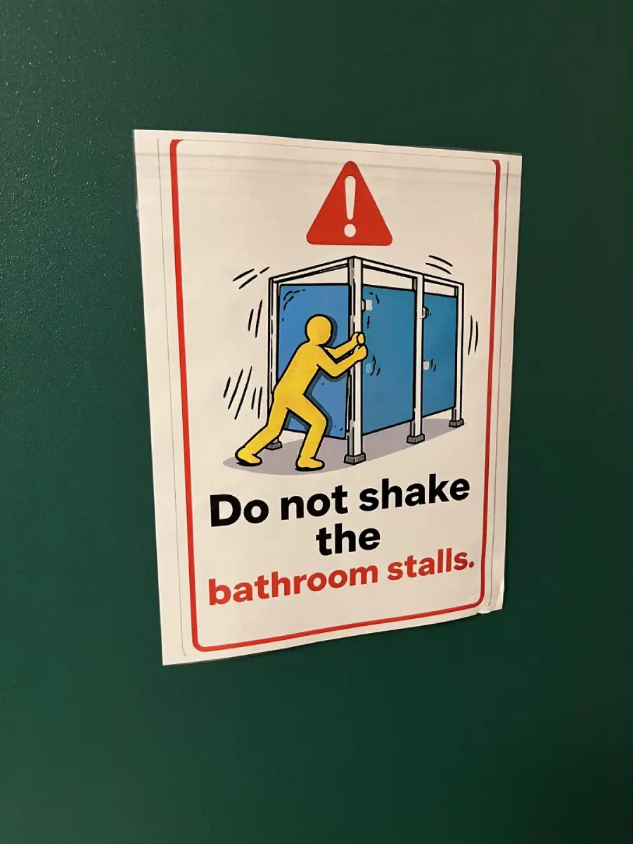 A sign on a green wall shows a yellow figure shaking a bathroom stall, with a red warning triangle above. Text reads: "Do not shake the bathroom stalls.