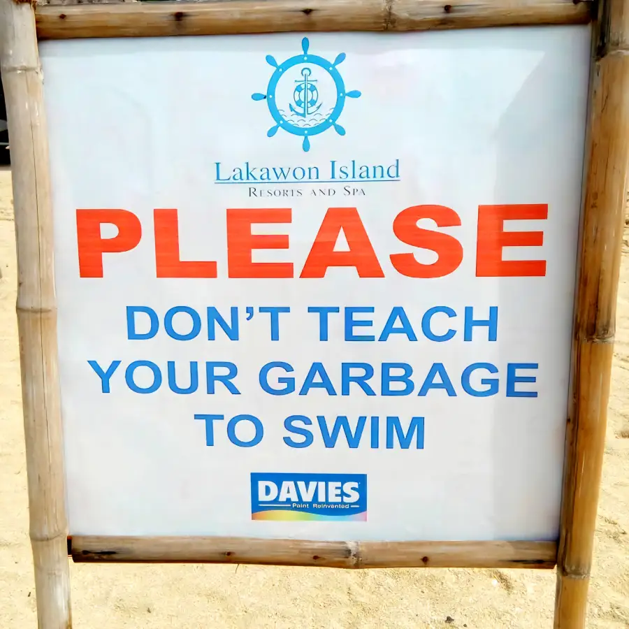 A beach sign reads: "Lakawon Island Resorts and Spa. PLEASE DON’T TEACH YOUR GARBAGE TO SWIM. DAVIES Paint Reinvented." The sign is bordered by bamboo and stands on sandy ground.