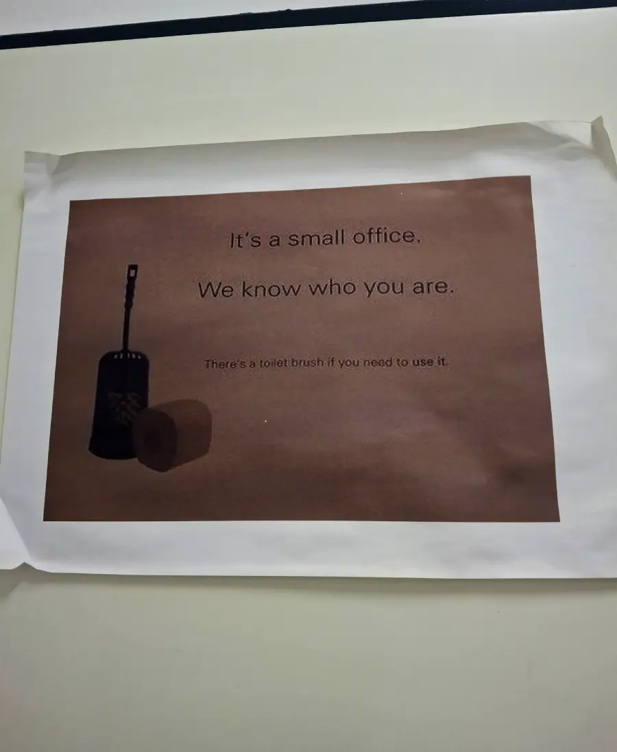 A printed sign on a wall reads: "It's a small office. We know who you are. There's a toilet brush if you need to use it." Next to the text are images of a toilet brush and a toilet paper roll.