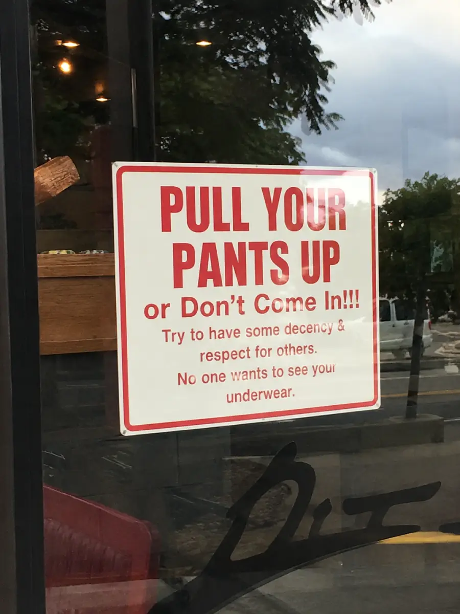 A sign on a glass door reads: “PULL YOUR PANTS UP or Don’t Come In!!! Try to have some decency & respect for others. No one wants to see your underwear.” Trees and a street are visible outside.