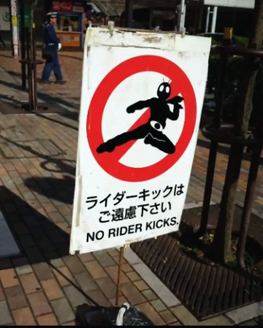 A sign with a silhouette of a masked superhero performing a flying kick inside a red prohibition circle. Text below reads, “NO RIDER KICKS” in English and Japanese. The sign is on a sidewalk.