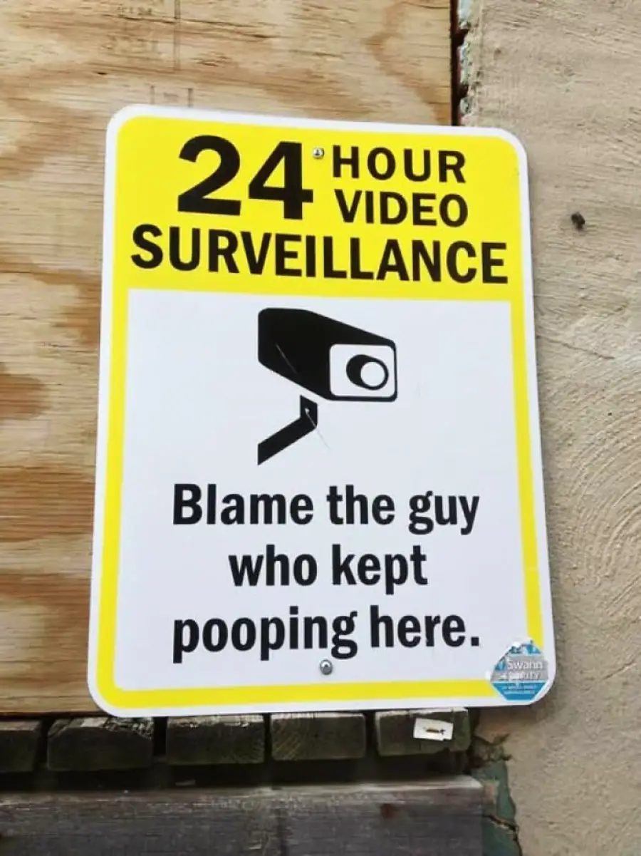 A sign reads "24 hour video surveillance" with a camera icon, and below it says, "Blame the guy who kept pooping here." The sign is attached to a wooden and concrete wall.