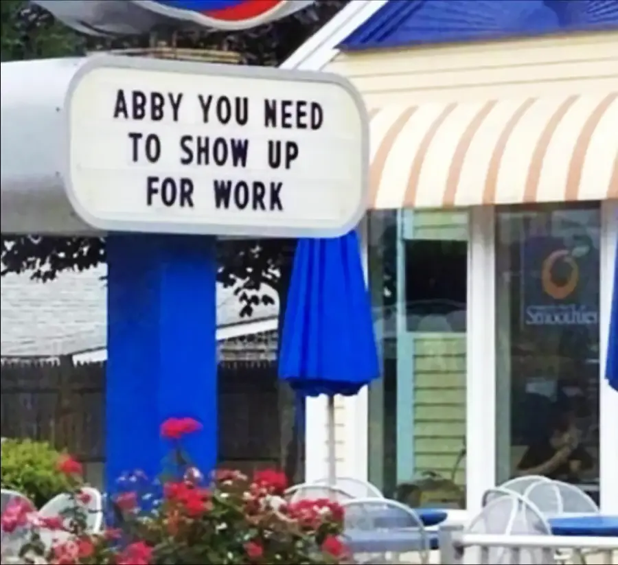 A restaurant sign outside a building reads, “ABBY YOU NEED TO SHOW UP FOR WORK,” above a patio with tables, chairs, flowers, and a blue umbrella.