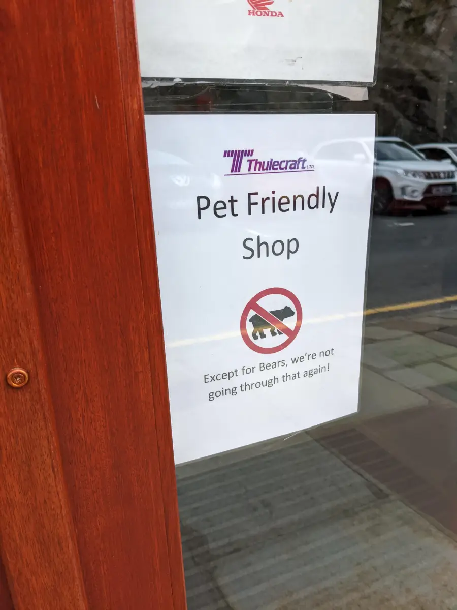 A sign on a shop door reads: "Pet Friendly Shop" with a symbol of a bear crossed out. Text below says, "Except for Bears, we're not going through that again!" Cars are visible reflected in the glass.
