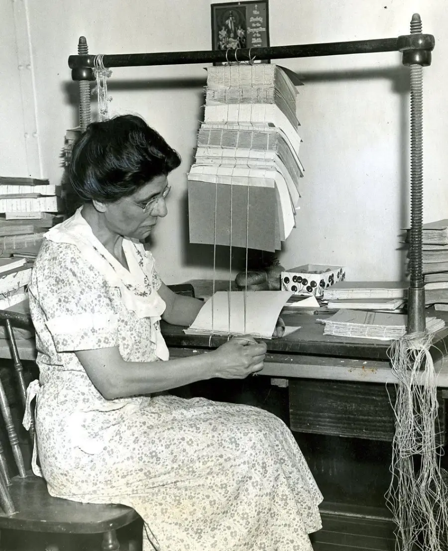 A woman in a patterned dress sits at a table, hand-sewing pages with thread using a bookbinding frame. Books and papers are stacked nearby.