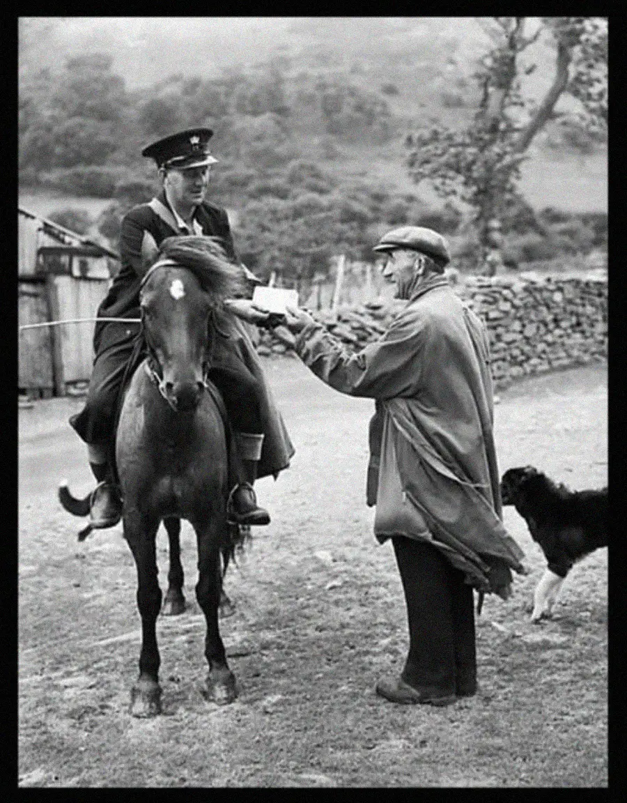 A mail carrier on horseback hands a letter to an older man in a coat, with a dog standing nearby and a rural landscape in the background.