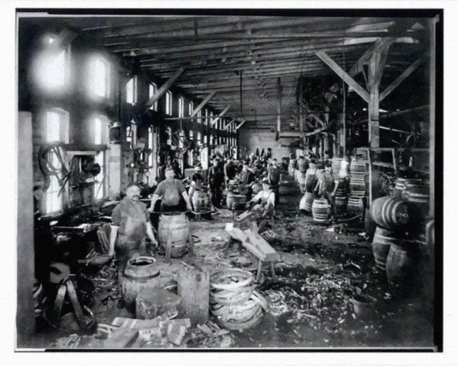 Black-and-white photo of workers in a large workshop assembling wooden barrels. Wooden beams, tools, and finished barrels are visible, with wood shavings scattered on the floor. Light streams in through the windows.