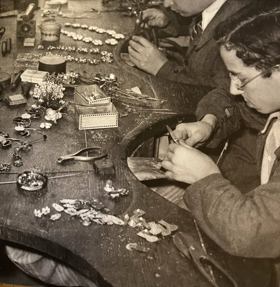 Two people work at a cluttered table assembling small jewelry pieces, with tools and decorative items spread out in front of them. They are focused on detailed, delicate craftwork.