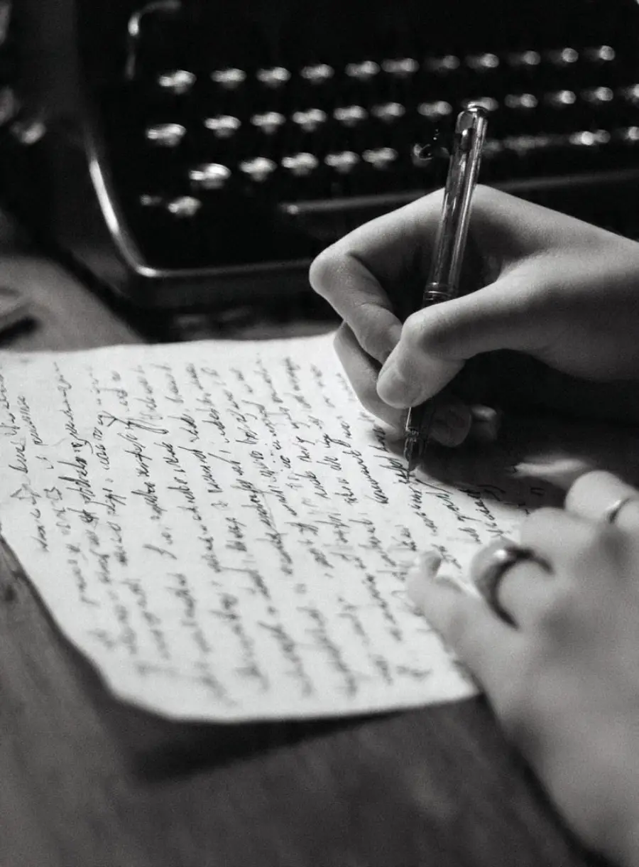 A person writes with a pen on a sheet of handwritten paper, with a typewriter visible in the background on a wooden desk.