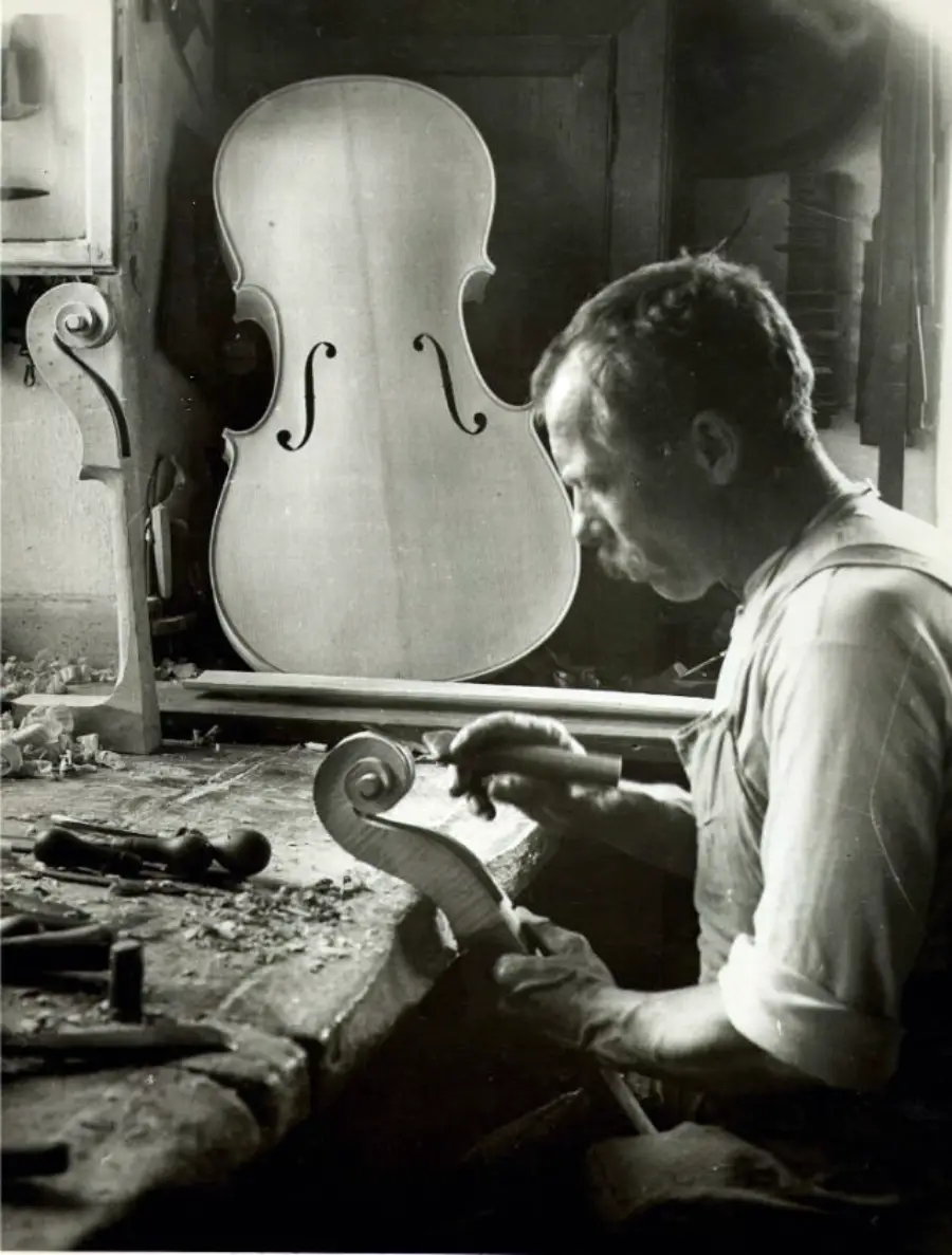 A luthier carves a cello scroll at a workbench, surrounded by woodworking tools, with an unfinished cello body propped up in the background.