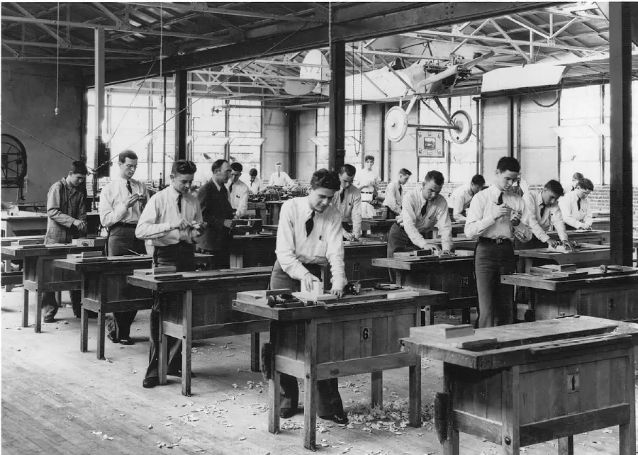A group of young men work at wooden benches in a spacious workshop, focusing on woodworking tasks. Sunlight streams through large windows, and a flying machine model hangs from the ceiling above them.