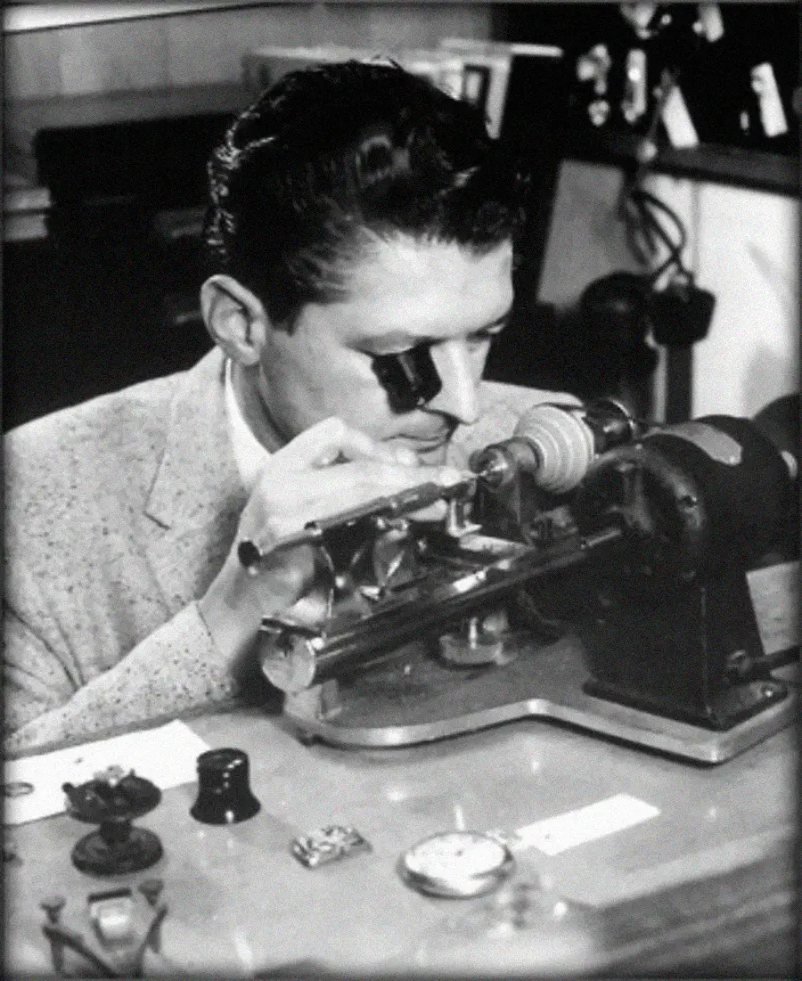 A man uses a monocle loupe to closely examine or repair a watch mechanism at a workbench with various tools and watch parts.