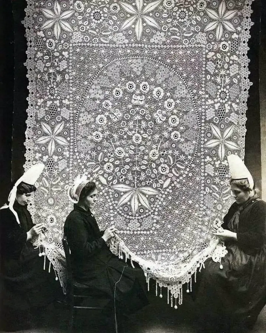 Three women in traditional clothing and tall hats sit in front of an elaborate, large lace textile, carefully working on the intricate design with their hands. The lace features detailed floral and star patterns.