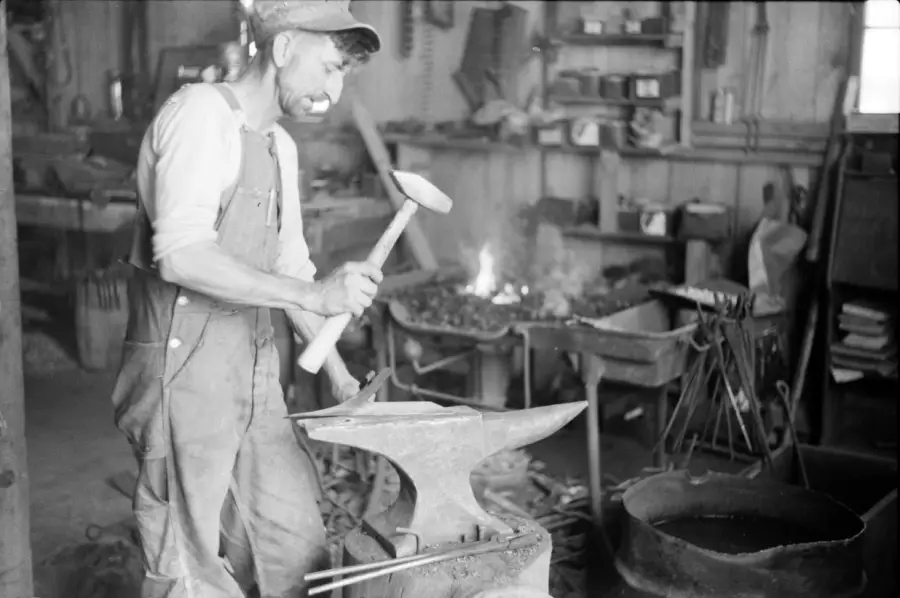 A blacksmith in overalls and a cap hammers hot metal on an anvil in a workshop, with tools and a forge fire visible in the background.