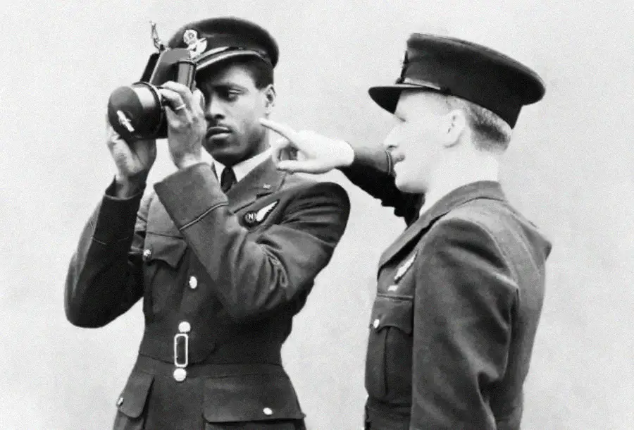 Two men in military uniforms from the mid-20th century, one looking through a camera while the other gestures and smiles, standing close together against a plain background.