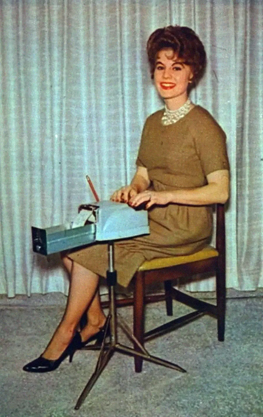A woman in a brown dress and black heels sits on a chair, typing on a manual typewriter with a pencil beside her. She is smiling, and there are white curtains in the background.