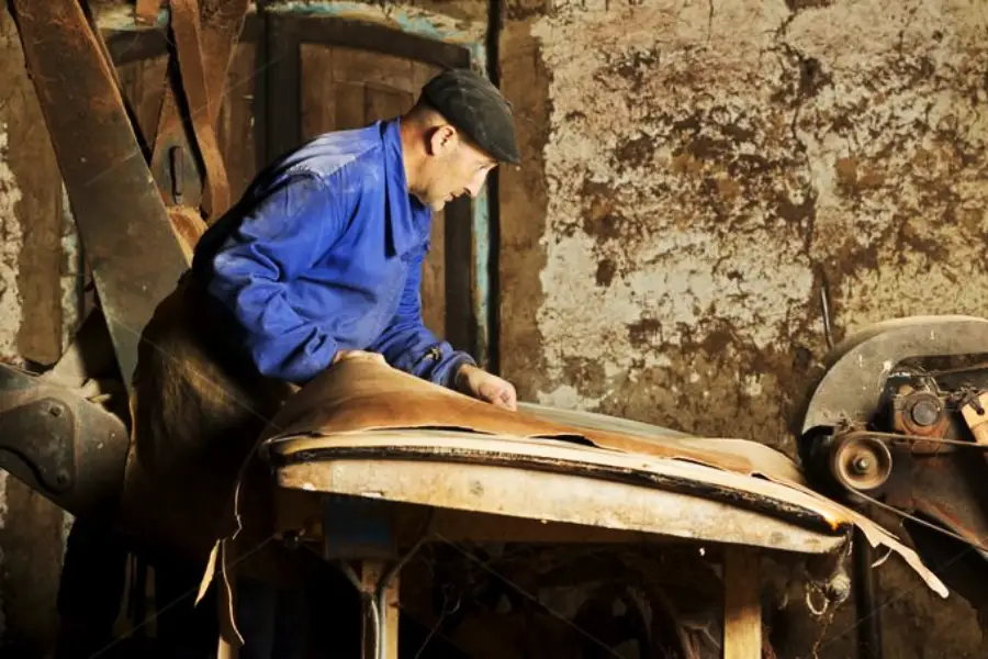 A man in a blue jacket and cap works with leather on a worn wooden table in a rustic, dimly lit workshop with aged, textured walls and old machinery.