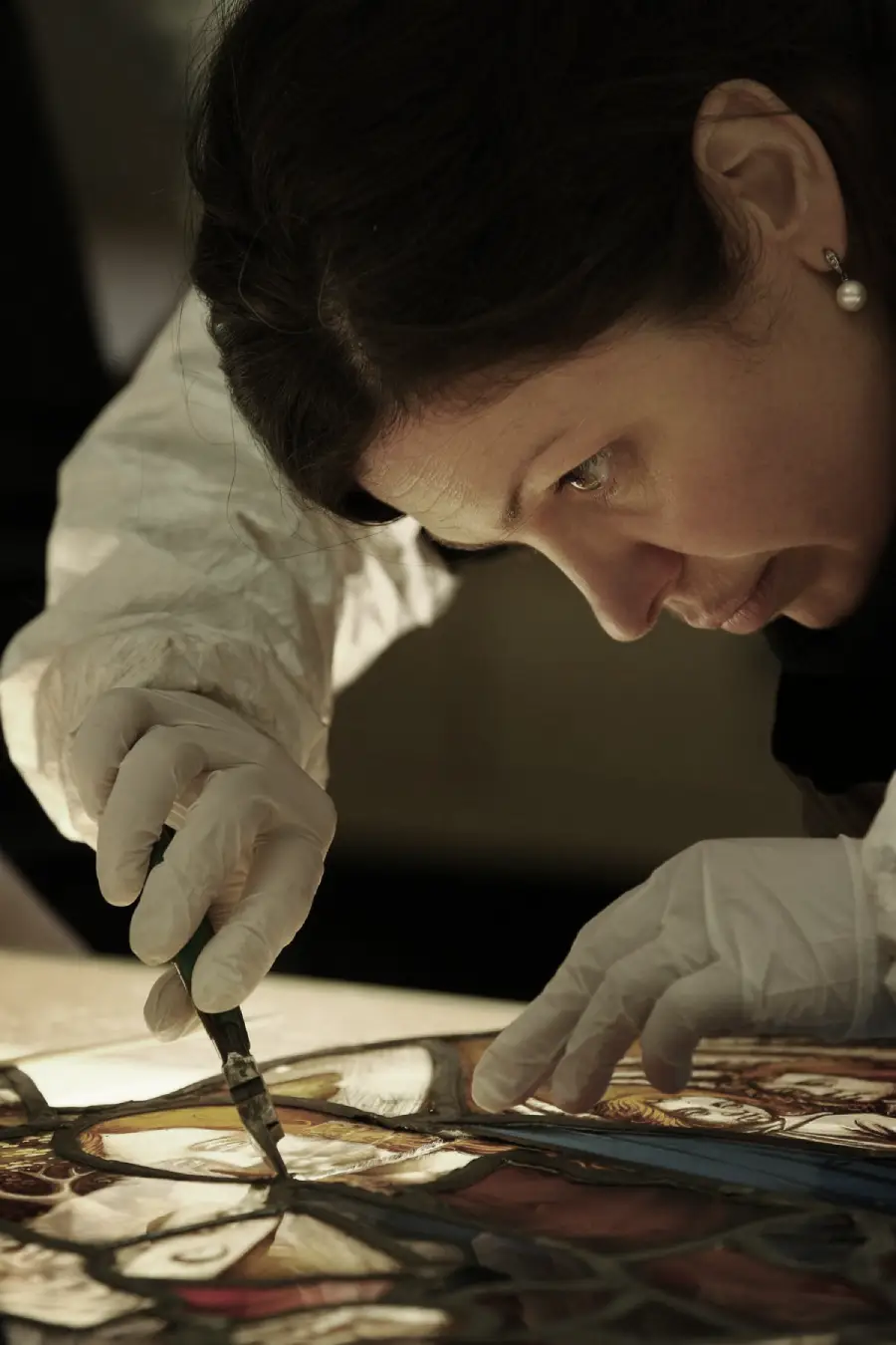 A woman wearing white gloves and a lab coat carefully restores a stained glass artwork using a brush, focusing intently on her work.