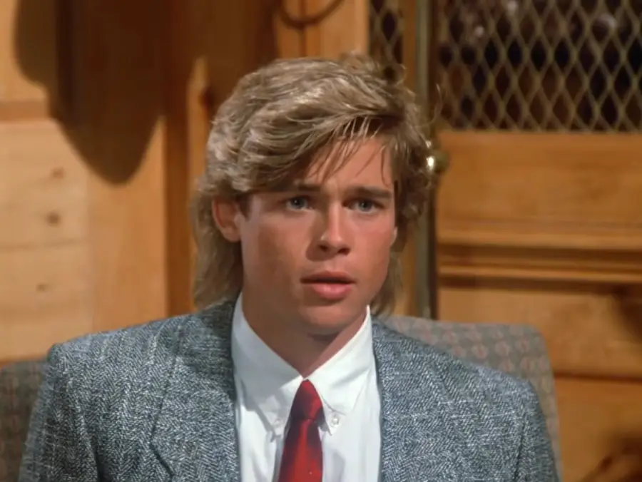 A young man with blond hair wearing a gray suit jacket, white shirt, and red tie sits indoors, looking slightly surprised. Wood paneling and a mesh-covered cabinet are in the background.