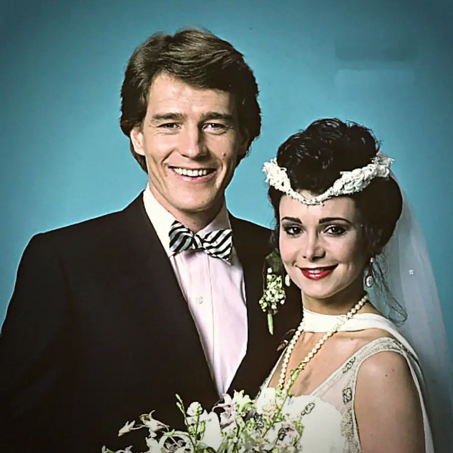 A smiling couple poses for a wedding portrait against a blue background. The man wears a black tuxedo and bow tie; the woman wears a white dress, veil, and floral headpiece, holding a bouquet of flowers.