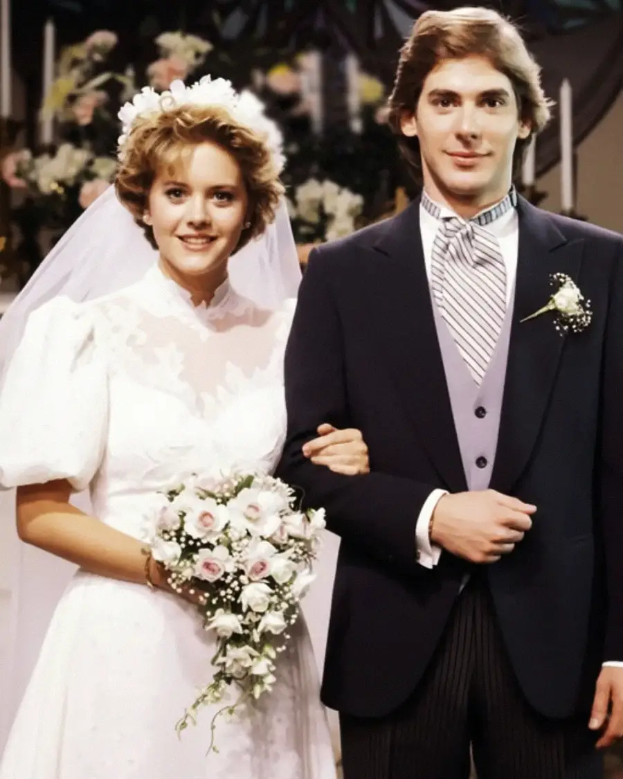 A bride in a white wedding dress and veil holds a bouquet, standing arm-in-arm with a groom in a dark suit and tie. They are posing and smiling in front of floral arrangements at an indoor wedding ceremony.
